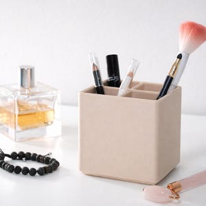 May include: A beige square makeup organizer holds various cosmetic items, including a makeup brush with a pink head, lip pencils, and a black lipstick tube. A clear perfume bottle, a black beaded bracelet, and a pink face roller are also visible.