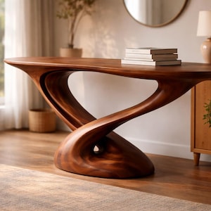 Op de afbeelding: Een handgemaakte, sculpturale houten console met een uniek, vloeiend ontwerp. Het rijke, donkerbruine hout heeft een gebogen basis en bovenkant, met een open, draaiende centrale steun. Er liggen verschillende boeken op het tafelblad. De tafel is een statement meubelstuk.