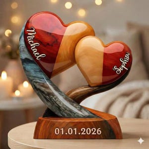 May include: A wooden heart-shaped sculpture with two connected hearts. One heart is red and brown, the other is orange and brown. The names "Michael" and "Sophia" are inscribed on the hearts. The base has the date "01.01.2026".