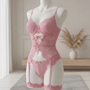 Puede incluir: Conjunto de lencería de encaje rosa sobre un maniquí. El conjunto incluye un bustier con cordones en la parte delantera, un tanga a juego y una liga. La lencería es de color rosa suave y presenta detalles de encaje floral.
