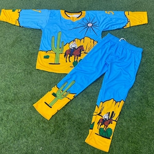 May include: A two-piece pajama set featuring a desert scene. The top is light blue with yellow accents, a V-neck, and long sleeves. The pants are light blue. The design includes cacti, a cowboy on a horse, and a sun graphic.