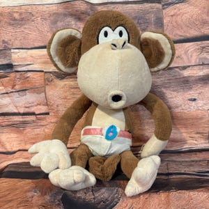 May include: A plush monkey toy with brown fur, a tan face, and white hands and feet. The monkey wears a white diaper with a pink belt and a blue accent. The toy is sitting on a wooden surface.