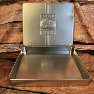 May include: Silver-colored rectangular tin with an open lid. The lid features the embossed word "FOSSIL" above a globe design and the number "2000". The tin is empty and sits on a wooden surface.