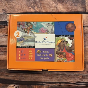 May include: An orange rectangular box for Hooked On Phonics Level 2. The front features illustrations of dolphins, a bear playing basketball, and text including 'Slam and Dunk are pals', 'workbooks books', 'audio tapes flash cards', and word lists.