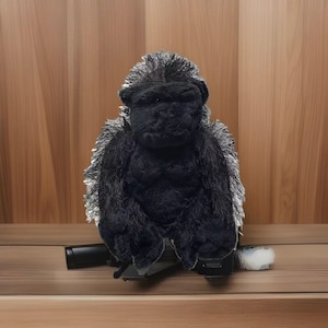 May include: A plush gorilla toy with black fur and gray accents. The gorilla is sitting on a black stand against a wooden background. The toy has a detailed face and is designed to resemble a real gorilla.
