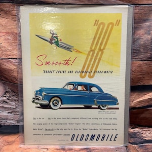 May include: A vintage Oldsmobile advertisement. The image shows a blue car with white tyres. The ad includes the text "Smoo-o-th!" and "Rocket Engine and Oldsmobile Hydra-Matic." A rocket ship graphic is also present, along with the year 1950.
