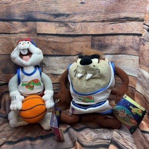 May include: Two plush toys of cartoon characters Bugs Bunny and Taz from Space Jam. Bugs Bunny is wearing a basketball jersey and holding a basketball. Taz is wearing a basketball jersey. Both toys have tags attached.