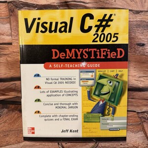 Puede incluir: Un libro titulado "Visual C# 2005 DeMYSTiFieD" con una cubierta amarilla y negra. La portada presenta un personaje de dibujos animados en una pantalla de ordenador y texto que destaca el formato de guía de autoaprendizaje y el contenido del libro.