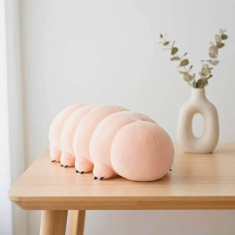 Puede incluir: Un coj&iacute;n suave en forma de oruga rosa con pies negros descansa sobre una mesa de madera clara. Los segmentos redondeados del coj&iacute;n crean un dise&ntilde;o caprichoso. Un jarr&oacute;n blanco con un recorte &uacute;nico y follaje seco est&aacute; en el fondo.