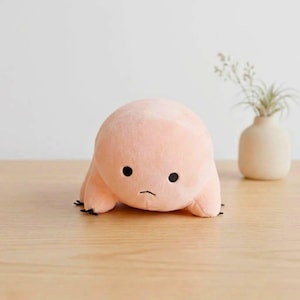 Puede incluir: Un peluche de color melocot&oacute;n en forma de oso de agua, con ojos negros y peque&ntilde;as garras negras. El juguete de peluche est&aacute; sobre una superficie de madera clara. Un peque&ntilde;o jarr&oacute;n con vegetaci&oacute;n est&aacute; en el fondo.