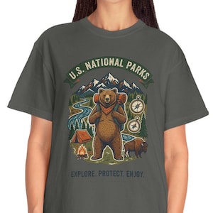 Puede incluir: Camiseta gris oscuro con un gráfico de un oso con una mochila, montañas y una brújula. El texto dice "U.S. National Parks. Explore. Protect. Enjoy."