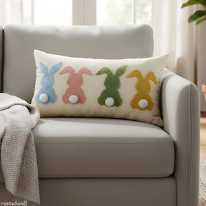 Könnte beinhalten: Ein rechteckiges Zierkissen mit vier bunten Hasenmotiven. Die Hasen sind blau, rosa, grün und gelb, jeder mit einem weißen Bommel-Schwanz. Das Kissen liegt auf einem grauen Sofa.
