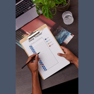 Op de afbeelding: Een houten clipboard met een "Sleep Tracker"-diagram wordt vastgehouden. De grafiek heeft een raster voor het volgen van slaappatronen, met kleurgecodeerde secties en een pen in de hand. Een laptop en plant staan op de achtergrond.