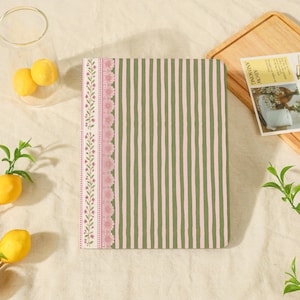 May include: A rectangular notebook featuring a cover with vertical olive green and light pink stripes, accented by a vertical border on the left side decorated with a delicate pink floral vine pattern and a textured pink scalloped edge detail.