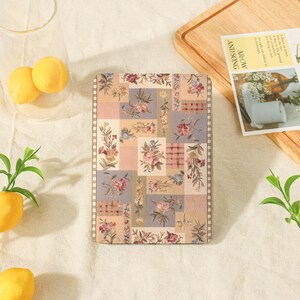 May include: A rectangular e-reader case featuring a patchwork design of floral patterns in muted shades of pink, blue, green, and beige. The cover has a checkered border and a textured surface, displaying various botanical illustrations throughout.