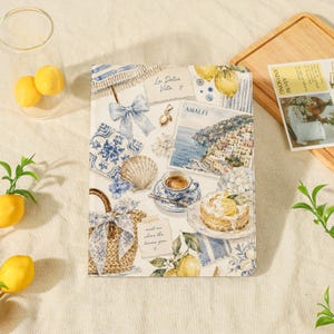 Könnte beinhalten: Ein rechteckiges Notizbuch mit einer Collage aus Amalfi-Illustrationen, darunter eine Küstenstadt, eine Tasse Kaffee, ein Zitronenkuchen, ein geflochtener Korb, eine Muschel und blaue Blumenmuster mit dem Text La Dolce Vita und Amalfi.