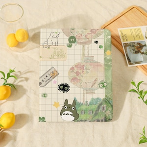 Op de afbeelding: Een rechthoekig notitieboek met een ruitjesachtergrond en diverse illustraties, waaronder een grote bloemenlantaarn, een groen huis, een klein grijs wezen en een label met de tekst KIKI'S Delivery Service in een collage-stijl.