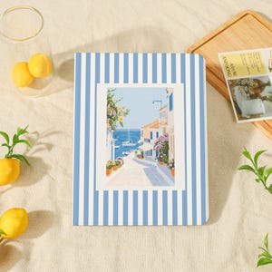 May include: A rectangular notebook featuring a blue and white vertical striped cover with a central illustration of a coastal Mediterranean street scene, including stone buildings, potted flowers, and a view of the blue sea with sailboats.