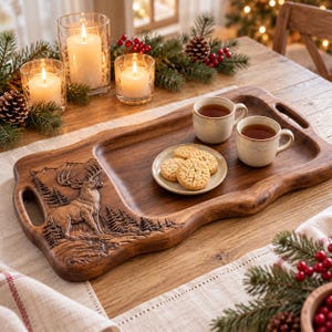 May include: A rectangular wooden serving tray with integrated handles, featuring a carved relief of a stag standing in a forest. The tray holds two ceramic mugs filled with dark liquid and a small plate containing three round, textured cookies.