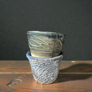 May include: Two handmade ceramic cups stacked on a wooden surface. The top cup is a muted blue-gray, while the bottom cup is a lighter blue. Both cups have a textured, organic design.