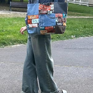 Peut inclure: Un sac fourre-tout fabriqué en tissu denim bleu avec un motif patchwork composé de diverses plaques d'immatriculation colorées. Le sac comprend deux bandoulières et un détail de poche visible sur le devant, affichant plusieurs codes alphanumériques.