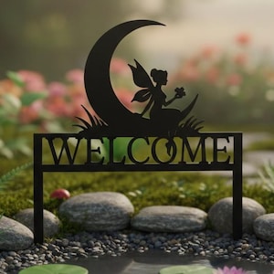 Puede incluir: Letrero de jardín de metal negro con un hada sobre una luna creciente y la palabra "WELCOME". El letrero está en un jardín con un estanque, rocas y flores rosas. Un elemento decorativo para exteriores.