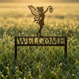 Könnte beinhalten: Dunkelbraunes Metall-Gartenschild mit dem Wort „WELCOME“ und einer Feen-Silhouette, die eine Laterne hält. Das Schild steht in einem Feld mit grünem Gras, mit der Fee oben.