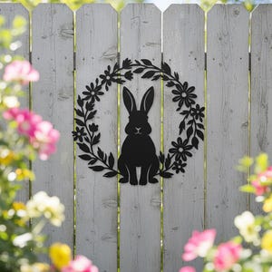 May include: A black metal rabbit silhouette within a floral wreath, mounted on a white wooden fence. The rabbit is centered, with long ears and a small tail. The wreath features leaves and flowers. Pink and yellow flowers are in the foreground.
