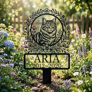 Könnte beinhalten: Schwarzer Metall-Gartenstecker mit einem Katzenporträt in einem floralen Kranz. Der Name "ARIA" und die Jahreszahlen "2012-2020" sind darunter abgebildet. Der Stecker ist für Gärten mit bunten Blumen gedacht.
