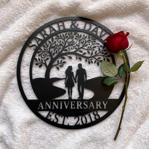 Könnte beinhalten: Ein rundes Wandbild aus schwarzem Metall mit der Silhouette eines Paares unter einem Baum. Der Text lautet SARAH & DAVID, ANNIVERSARY und EST. 2018. Eine einzelne rote Rose mit grünen Blättern liegt an der rechten Seite des Schildes.