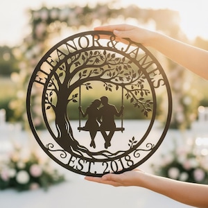 Könnte beinhalten: Ein kreisförmiges Wandbild aus dunkelbraunem Metall mit der Silhouette eines Paares auf einer Schaukel unter einem Baum. Der Text "ELEANOR & JAMES" ist oben gebogen und "EST. 2018" ist am unteren Rand des kreisförmigen Rahmens zentriert.