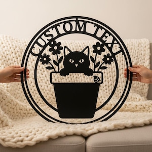 Könnte beinhalten: Schwarze Metall-Wandkunst mit einer Katze, die aus einem Blumentopf schaut, umgeben von Blumen und den Worten "CUSTOM TEXT" in einem kreisförmigen Design. Ein Marienkäfer ist ebenfalls vorhanden. Die Kunst wird von zwei Händen gehalten.