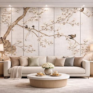 May include: A living room scene featuring a cream-colored sofa with several throw pillows, a round coffee table, and two side tables with lamps. The focal point is a large wall mural depicting a flowering tree with birds. The room has a neutral color palette.