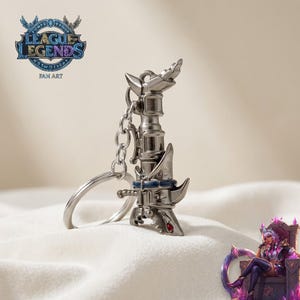 Op de afbeelding: Een zilverkleurige sleutelhanger met een gestileerd ontwerp in de vorm van een zwaard, een ketting en een sleutelring. De sleutelhanger heeft blauwe en rode accenten. Het League of Legends-logo is zichtbaar op de achtergrond.