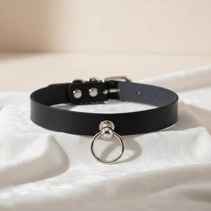 Op de afbeelding: Een zwarte leren halsband met een zilveren ring en gespsluiting. De halsband wordt gepresenteerd op een witte, getextureerde ondergrond. Een eenvoudig, maar stijlvol accessoire.