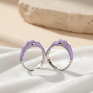 Peut inclure: Deux bagues assorties avec un motif en forme de goutte de couleur violet clair et une bande argentée. Les bagues ont un design minimaliste avec une petite bouche et deux points pour les yeux. Le bas des bagues est argenté.