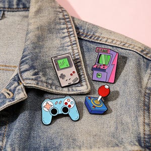 May include: Four enamel pins with a retro video game theme. Includes a gray handheld console, a purple arcade cabinet with "GAME" text, a blue controller, and a blue joystick with a red ball.