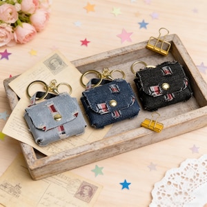 Puede incluir: Tres llaveros de mini bolsos estilo denim en azul claro, azul oscuro y negro. Cada uno tiene un cierre dorado y un asa pequeña. Los bolsos tienen detalles en rojo y blanco y un cierre de botón dorado. Se exhiben en una bandeja de madera con postales vintage.