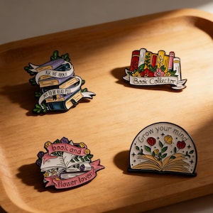Pode incluir: Quatro pinos de esmalte em uma bandeja de madeira. Os pinos apresentam designs com tema de livro com textos como "Ask Me About My To Be Read Pile" e "Book Collector". Outros pinos dizem "book and flower lover" e "Grow your mind".