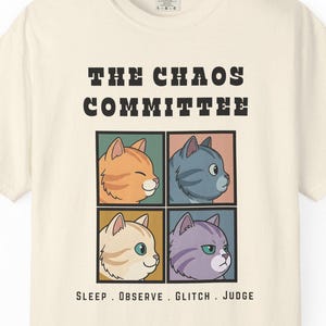 Op de afbeelding: Crèmekleurig T-shirt met de tekst "THE CHAOS COMMITTEE" boven vier cartoon kattengezichten in gekleurde vierkanten. Daaronder staan de woorden "SLEEP . OBSERVE . GLITCH . JUDGE". De katten hebben verschillende kleuren en uitdrukkingen.