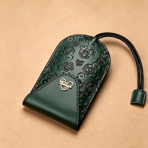 Puede incluir: Llavero de cuero verde oscuro con un patrón de Mickey Mouse en relieve. El llavero tiene un pequeño emblema dorado de Mickey Mouse y un cordón negro con una pequeña lengüeta verde.