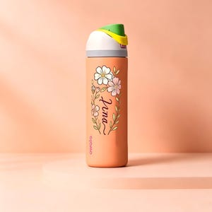 May include: An orange Owala water bottle featuring a floral design and the name Nina in black script. The bottle has a white lid with a green and yellow flip-top cap. The brand name Owala is printed in small pink letters near the bottom of the bottle.
