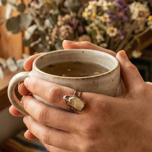 May include: A person holding a ceramic mug filled with tea. The mug is light-colored with a handle and is held near a window with natural light. A ring with a large, light-colored stone is on the person's finger. Dried flowers are in the background.
