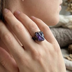 Peut inclure: Une bague en métal foncé ornée d'une améthyste violette brute de forme irrégulière. La bague est portée au doigt, la pierre précieuse étant bien en évidence. L'image montre également de délicates boucles d'oreilles couleur cuivre.