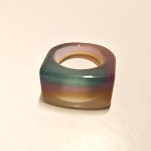 Op de afbeelding: Een vierkante ring met een kleurengradient van blauw, groen, paars en geel. De ring lijkt gemaakt van een doorschijnend materiaal, met een glad, gepolijst oppervlak. De ring is een modeaccessoire.