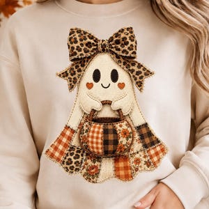 May include: A beige crewneck sweatshirt featuring a central graphic of a ghost with a leopard print bow. The ghost holds a pumpkin-shaped basket decorated with various autumn-themed patterns including plaid, floral, and leopard print designs.