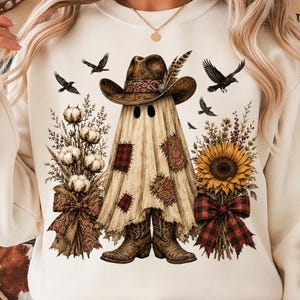 Op de afbeelding: Een crèmekleurige sweater met ronde hals en een afbeelding van een geest die een cowboyhoed en laarzen draagt. De geest is omringd door katoentakken, een zonnebloem, vliegende kraaien en stoffen lapjes met een ruitpatroon.