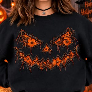 Könnte beinhalten: Ein schwarzes Langarm-Sweatshirt mit einer großen, stilisierten Kürbisgesicht-Grafik auf der Vorderseite. Das Design besteht aus orangefarbenen Spitzenstoff-Applikationen mit ausgefransten Rändern, die Augen, Nase und ein gezacktes Grinsen bilden.