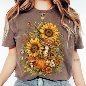 Può includere: Una t-shirt marrone a maniche corte con una stampa grafica raffigurante due grandi girasoli gialli, una zucca arancione centrale e due funghi rossi a pois bianchi circondati da varie foglie verdi e fiori più piccoli arancioni e bianchi.