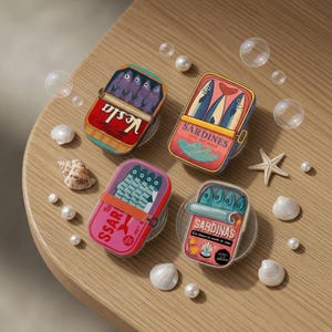 Puede incluir: Cuatro agarres para teléfono con temática de sardinas con ilustraciones coloridas de peces enlatados. Los agarres están dispuestos sobre una superficie de madera con conchas marinas, perlas y burbujas. Cada agarre presenta un diseño diferente de lata de sardinas.